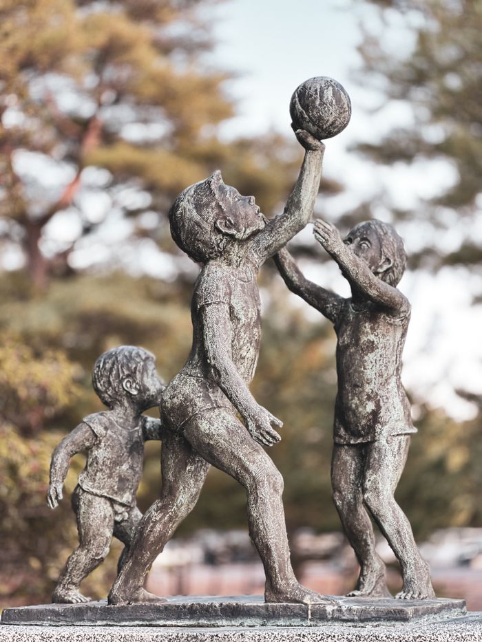 Bronze statue depicting three children playing with a ball. Captured outdoors.