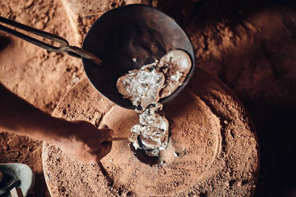 A foundry worker pours molten metal from a crucible onto sand, showcasing traditional smelting techniques.